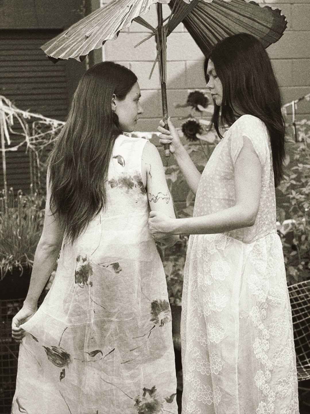 Two women in long white dresses standing outdoors under a traditional umbrella. The image is in black and white, showing floral patterns on the dress on the left and lace detailing on the dress on the right.